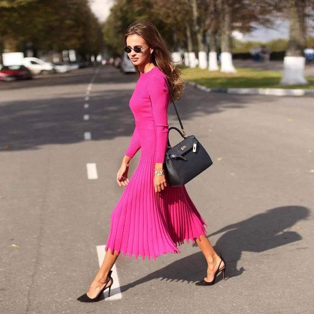 Robe plissée midi rose fluide manches longues pour cocktail chic femme, accessoirisée d’un sac noir élégant