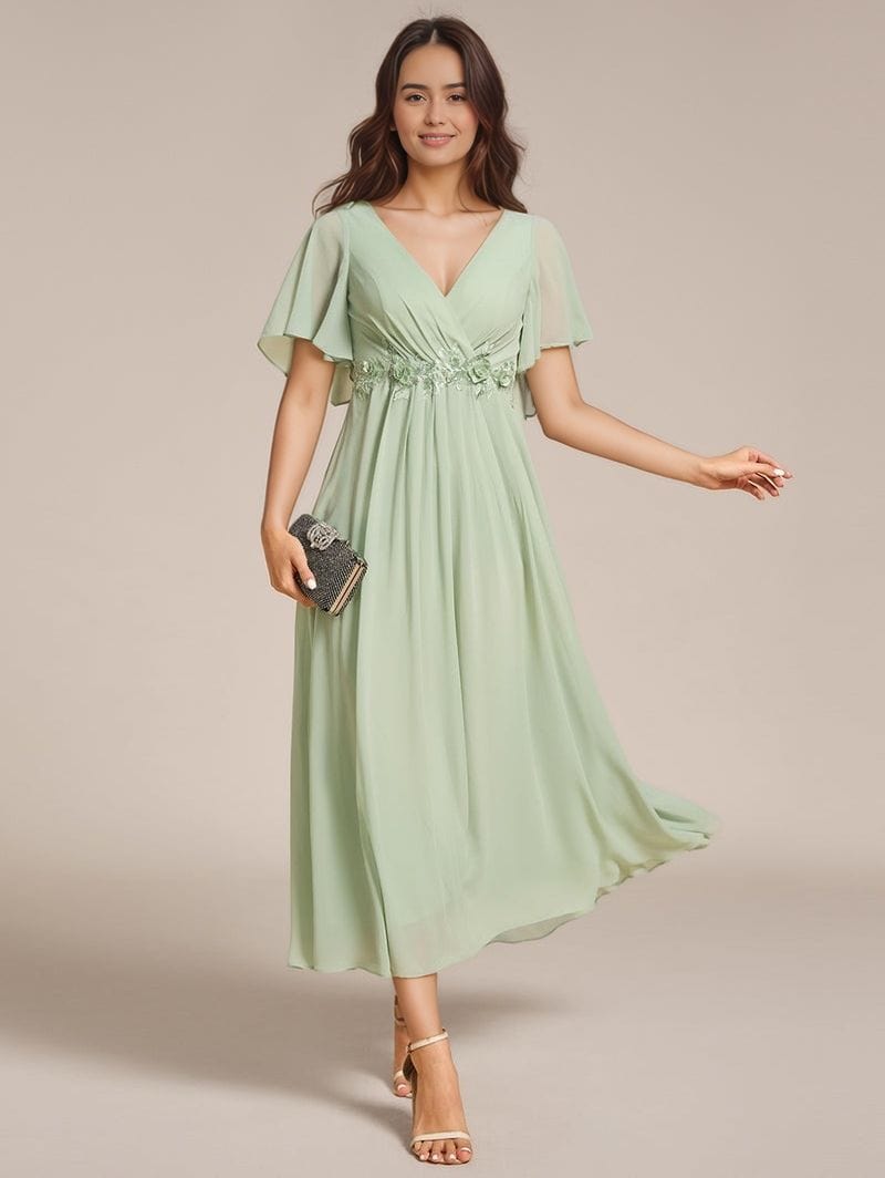 Robe de soirée bohème longue verte manches courtes fluide avec décolleté femme élégante tenant une pochette
